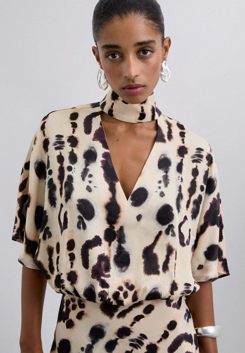 Mujer con el pelo peinado hacia atrás que lleva una blusa de patrón abstracto en beige, negro y púrpura con escote recortado, y pendientes y pulsera de aros plateados.