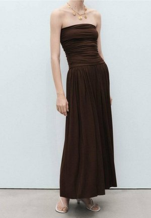 Donkerbruine strapless maxi-jurk met geplooide details, gedragen met witte open-toe hakken en gelaagde gouden muntenkettingen tegen een effen lichte muur.