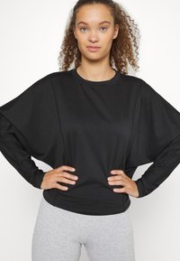 Top preto de mangas longas com mangas largas e caídas, e decote redondo, combinado com calças justas cinza claro. Textura de tecido suave.