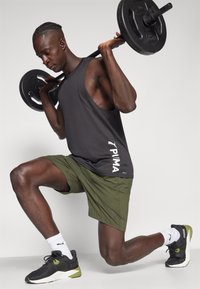 Débardeur noir avec de larges emmanchures, shorts verts, et baskets noires avec des accents olive, soulevant des poids en position à genoux.