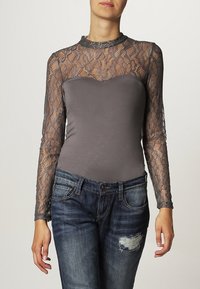 Grå långärmad topp med spetsöverlägg. Kroppen är figurnära, medan ärmarna har intrikata spetsdetaljer. Matchas med slitna blå jeans.