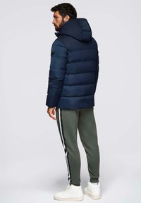 Giacca imbottita blu navy con cuciture orizzontali e cappuccio. Indossata sopra pantaloni verde oliva a sigaretta con strisce laterali e sneakers bianche.