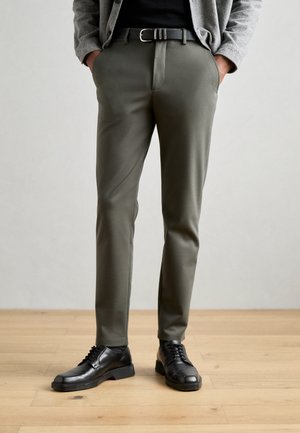 Slim-fit olive green trousers with a smooth texture, paired with black leather shoes. The outfit is complemented by a black belt and grey cardigan.