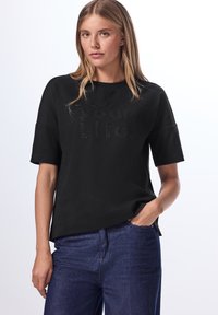 Top negro de manga corta con cuello redondo y texto en relieve "Vive tu vida". Diseñado con un corte relajado y una textura de tela suave.