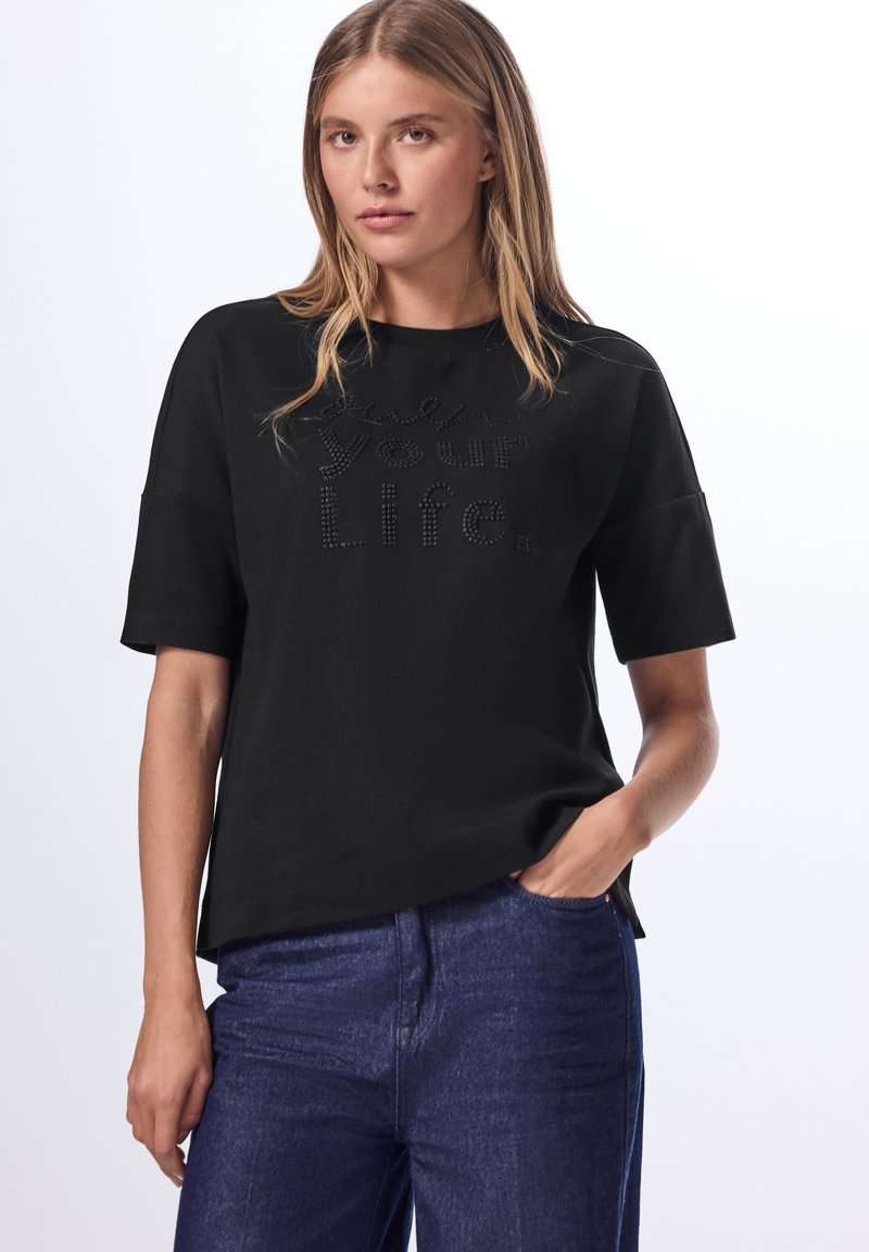 Top negro de manga corta con cuello redondo y texto en relieve "Vive tu vida". Diseñado con un corte relajado y una textura de tela suave.