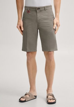 Mann trägt olivgrüne knielange Shorts mit Knopf- und Reißverschluss, kombiniert mit braunen Sandalen mit Schnallen und einem weißen Hemd.