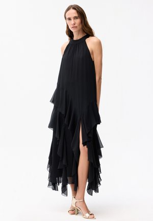 Frau trägt ein ärmelloses, schwarzes, asymmetrisches Kleid mit gestuften Rüschen und Seitenschlitz, kombiniert mit goldenen Sandalen mit hohen Absätzen.