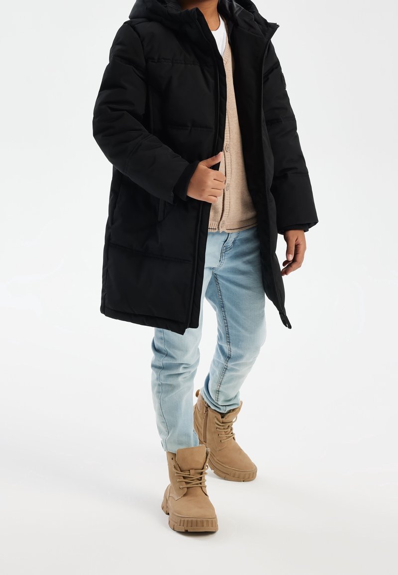 Enfant portant un jean bleu clair, des bottines marron clair à lacets, un cardigan beige, une chemise blanche et un manteau matelassé noir à capuche sur fond blanc.