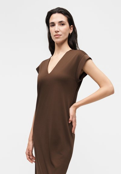 Γυναίκα με σκουρόχρωμα μαλλιά που φοράει ένα καφέ αμάνικο φόρεμα με V λαιμόκοψη, ποζάροντας με το ένα χέρι στη μέση σε απλό λευκό φόντο.