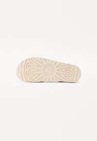 Såle av en sko i lys beige med teksturerte geometriske mønstre og et sirkulært emblem i midten. Glatt finish med et minimalistisk design.