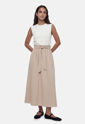 Vrouw staand met handen achter de rug, draagt een wit mouwloos topje, een beige hooggetailleerde gebonden rok en zwarte platte schoenen op witte achtergrond.