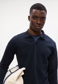 Tengeri kék, hosszú ujjú pólóing V-nyakkal, sima anyagból készült. A modell világos színű dzsekit tart, hangsúlyozva az öltözet kontrasztját.