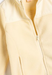 Jasnożółta bluza z polaru z gładką górną częścią, pełnym zapięciem na zamek i bocznymi kieszeniami. Materiał ma miękkie, teksturowane wykończenie.