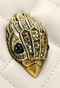 Złotona w bird-shaped hardware z czarnymi kamiennymi oczami i szczegółowym wzorem piór, na teksturowanym białym tle.