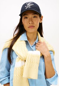 Gele gebreide trui gedrapeerd over een blauw gestreept overhemd, aangevuld met een marineblauwe pet met wit geborduurd logo. Soepele stoftexturen.