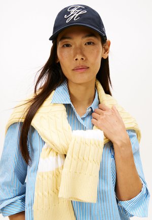 Gele gebreide trui gedrapeerd over een blauw gestreept overhemd, aangevuld met een marineblauwe pet met wit geborduurd logo. Soepele stoftexturen.