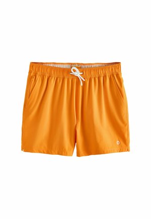 Muž bez trička, ktorý má na sebe oranžové plavky a slnečné okuliare, stojí bosý na skalnatom pobreží s morom a budovami na kopci v pozadí.