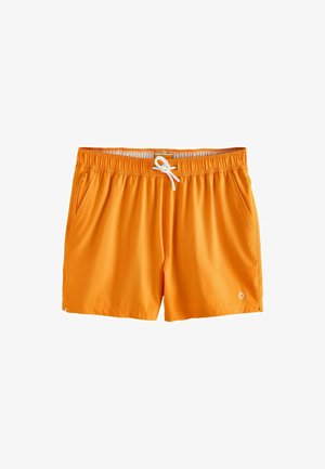 Bărbat fără tricou, purtând pantaloni de baie portocalii și ochelari de soare, stă desculț pe malul stâncos cu mare și clădiri pe deal în fundal.