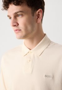 Beige polo póló texturált gallérral, két gombbal és hímzett "BOSS" logóval a mellkason. Simácska, könnyű anyag.