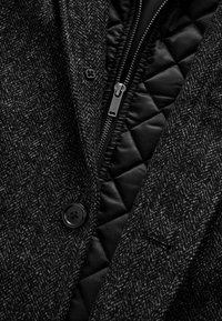 Jachetă din tweed negru cu un model de herringbone, având o căptușeală neagră matlasată și un fermoar argintiu, completată de un nasture negru.