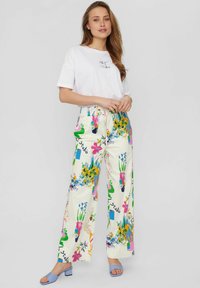 Weißes T-Shirt mit "ART Studio"-Logo, kombiniert mit cremefarbenen weiten Hosen mit bunten Blumenmustern und einem Kordelzugbund. Blaue Sandalen.
