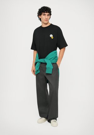 T-shirt nera con logo grafico e sorriso giallo, abbinata a pantaloni grigi larghi e un maglione teal legato in vita. Sneakers bianche.