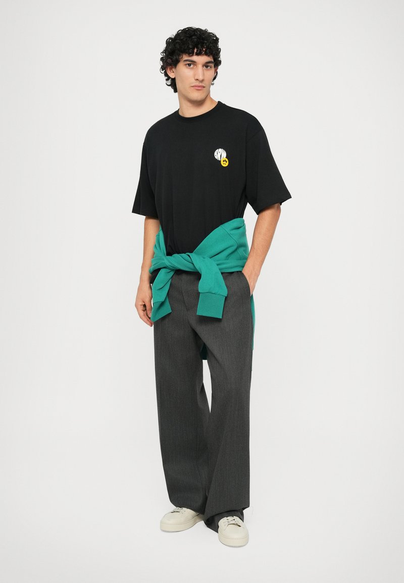 T-shirt nera con logo grafico e sorriso giallo, abbinata a pantaloni grigi larghi e un maglione teal legato in vita. Sneakers bianche.