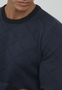 Navy sweater with a black ribbed neckline featuring a diamond pattern. The fabric has a soft texture and a fitted design.