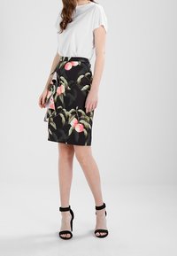 Žena v bílém topu a černé sukni s růžovým ovocem a zeleným listovým vzorem, doplněná černými sandály na vysokém podpatku.