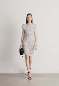 Vestido de lantejoulas prateadas com uma silhueta ajustada, ombros largos e detalhe franzido. Combinado com uma pequena bolsa preta e saltos pretos com tiras.