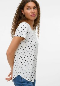 Hvid T-shirt med korte ærmer, marineblå polka-dots, rund halsudskæring og bølget kant. Fremstillet af let materiale, velegnet til afslappet brug.