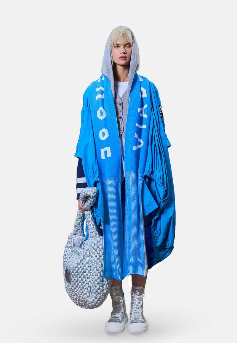 Personne portant un manteau bleu oversize superposé, un sweat à capuche gris, une longue écharpe bleue avec un texte blanc, des baskets montantes argentées, portant un grand sac texturé.