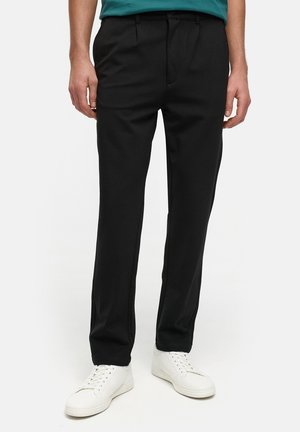 Schwarze maßgeschneiderte Hose mit glatter Textur, mit Vorderfalten und schmaler Passform. Kombiniert mit weißen Sneakers für einen Kontrast.