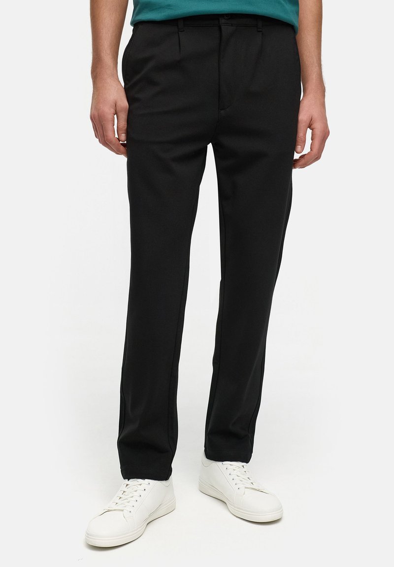 Pantaloni neri su misura con una texture liscia, dotati di pieghe frontali e una vestibilità slim. Abbinati a sneakers bianche per un contrasto.