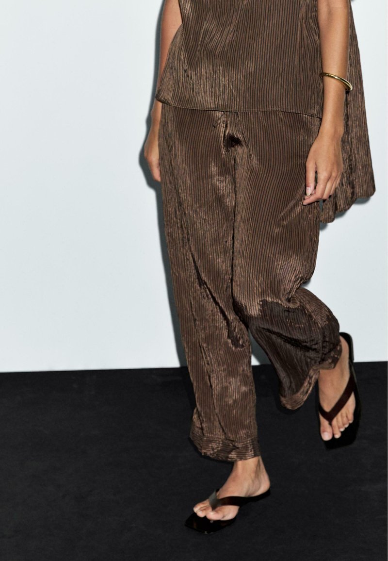 Top plissettato marrone e pantaloni abbinati, caratterizzati da una vestibilità comoda e una texture morbida. Sandali neri semplici completano il look.
