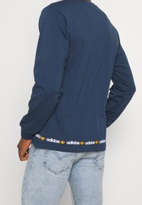 Námořnický modrý tričko s dlouhým rukávem vyrobené z bavlny s kulatým výstřihem, které má kontrastní bílou a oranžovou adidas logo pásku na lemu.