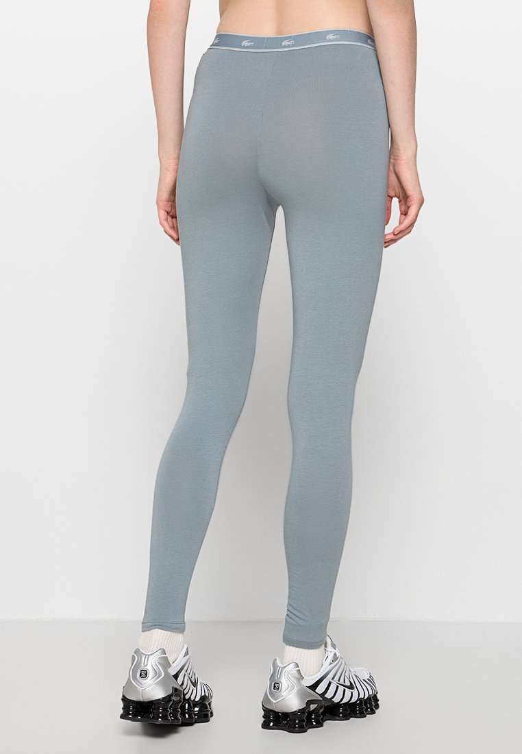 Leggings de color gris claro, hechos de un material suave y elástico con un diseño ajustado y una cinturilla en contraste. Presenta una textura sutil; combinados con zapatillas deportivas.