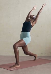 Persoon in een zwart topje en lichtblauwe shorts die een yoga halvemaanuitvalsoefening uitvoert op een roze mat tegen een effen muur.