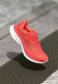 Helle korallenfarbene Laufenschuhe mit weißer Sohle, in der Luft über einer Asphaltstraße, die Seiten- und Obendetails einschließlich Schnürsenkeln und Logo zeigen.