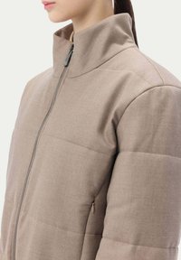 Vadderad beige jacka med hög krage, dragkedja fram och sidofickor. Tyget har en slät textur med ett quiltat mönster.