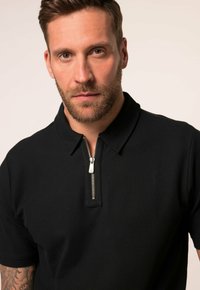 Černé polo tričko s límcem na zip, krátké rukávy, vyrobené z texturované látky. Obsahuje viditelný stříbrný zip.