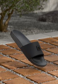 Sandelă neagră cu braț lat care prezintă un logo Hugo, plasată deasupra unei suprafețe cu dale de lemn, având mici pietricele albe între ele.