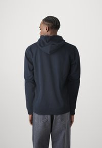 Felpa con cappuccio blu navy, dotata di tasca frontale a marsupio, polsini e orlo a coste. Presenta una texture morbida e una vestibilità comoda, abbinata a jeans grigi chiari.