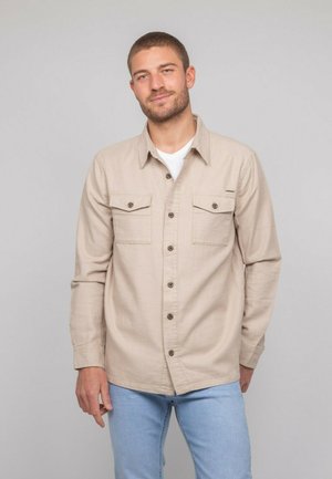 Chemise beige à manches longues, boutonnée, avec deux poches poitrine, boutons marron et coupe droite. Portée par-dessus un t-shirt blanc.