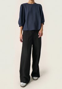 Osoba oblečená v námořnicky modrém tričku s nafouklými rukávy, širokých černých kalhotách a světlých šedých teniskách stojící na neutrálním pozadí.