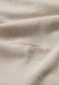 Bēša adīta auduma ar smalku tekstūru, maigu izskatu un nelielu stiepšanos. Iekļauta neliela redzama šuvju detaļa uz virsmas.