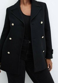 Abrigo negro de lana de doble botonadura con botones dorados, solapas con muescas y acabado texturizado. Combinado con vaqueros negros.