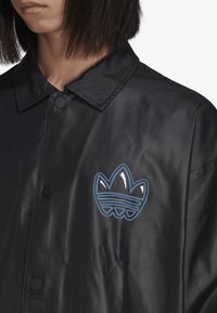 Veste noire, brillante, à boutonnière avec un col pointu. Présente un logo graphique bleu sur la poitrine gauche et des boutons noirs. Texture lisse.
