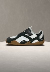 Chaussure de sport blanche et noire avec semelle marron, dessus en mesh et bande velcro, exposée sur une surface claire avec un fond uni.
