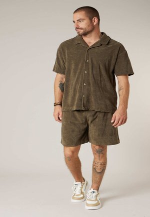 Homme tatoué portant une chemise à manches courtes boutonnée vert olive et un short assorti, debout avec des baskets blanches et beiges sur un fond neutre.